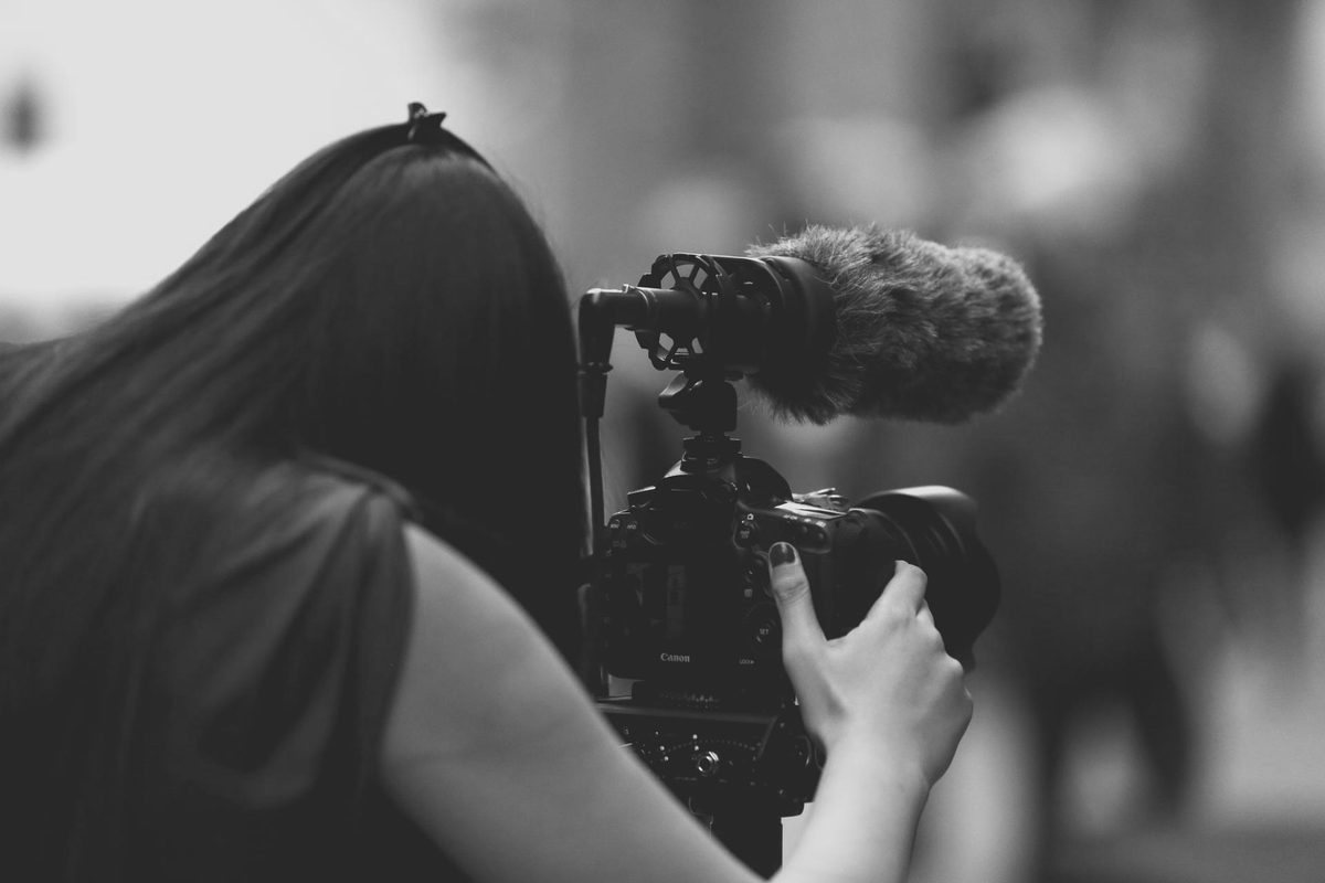 A camera operator filming, a shotgun microphone mounted on the camera